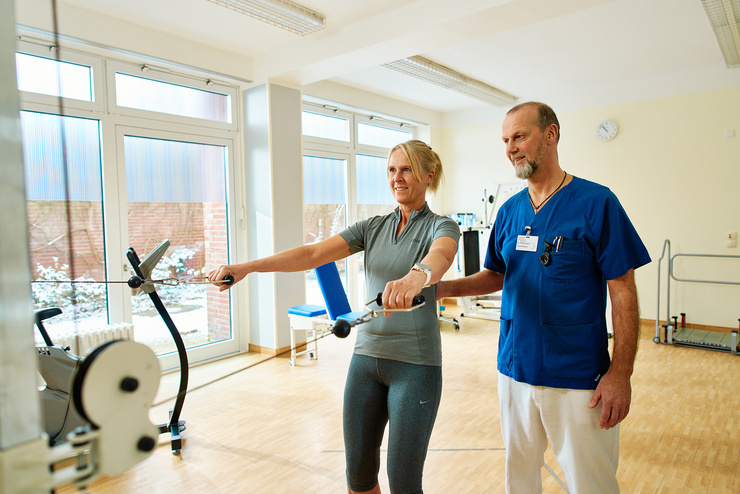 Blick in einen sonnigen Therapieraum, ein Physiotherapeut leitet eine Patientin an, während einen weitere Patientin im Hintergrund trainiert.