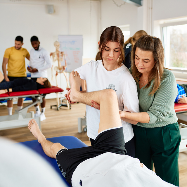 Praktischer Unterricht in der Physiotherapieschule. Eine Schülerin wird von einer Lehrerin angeleitet und führt eine Übung am rechten Bein eines Patienten, der auf einer Liege liegt. 